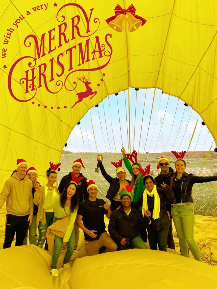 Group of people in Christmas hats inside a yellow hot air balloon with 'Merry Christmas' text.